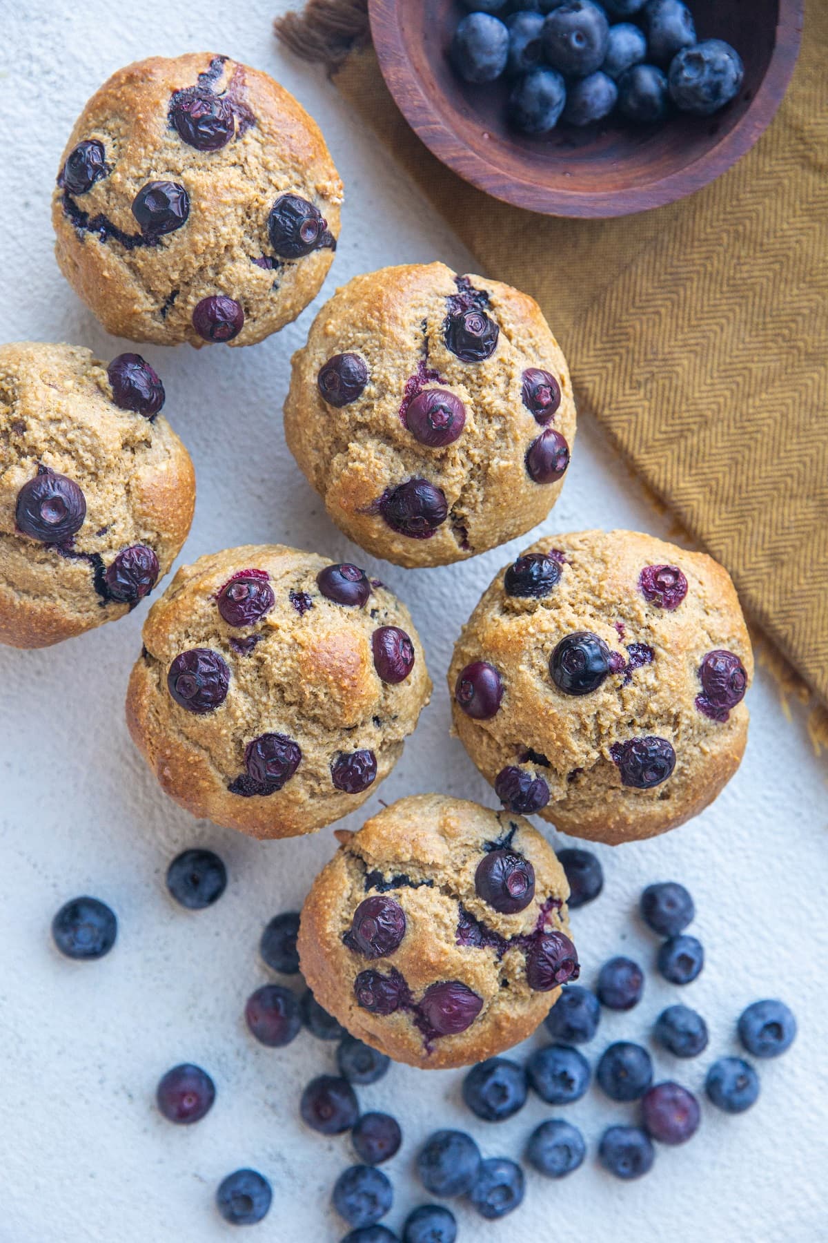 Six blueberry banana muffins on a white background with a golden napkin and fresh blueberries scattered about.