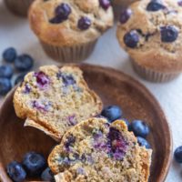 Plate with a muffin sliced in half and a hand picking up one of the halves, with full muffins in the background.