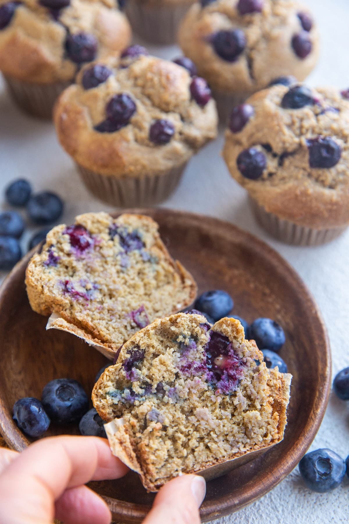 Plate with a muffin sliced in half and a hand picking up one of the halves, with full muffins in the background.