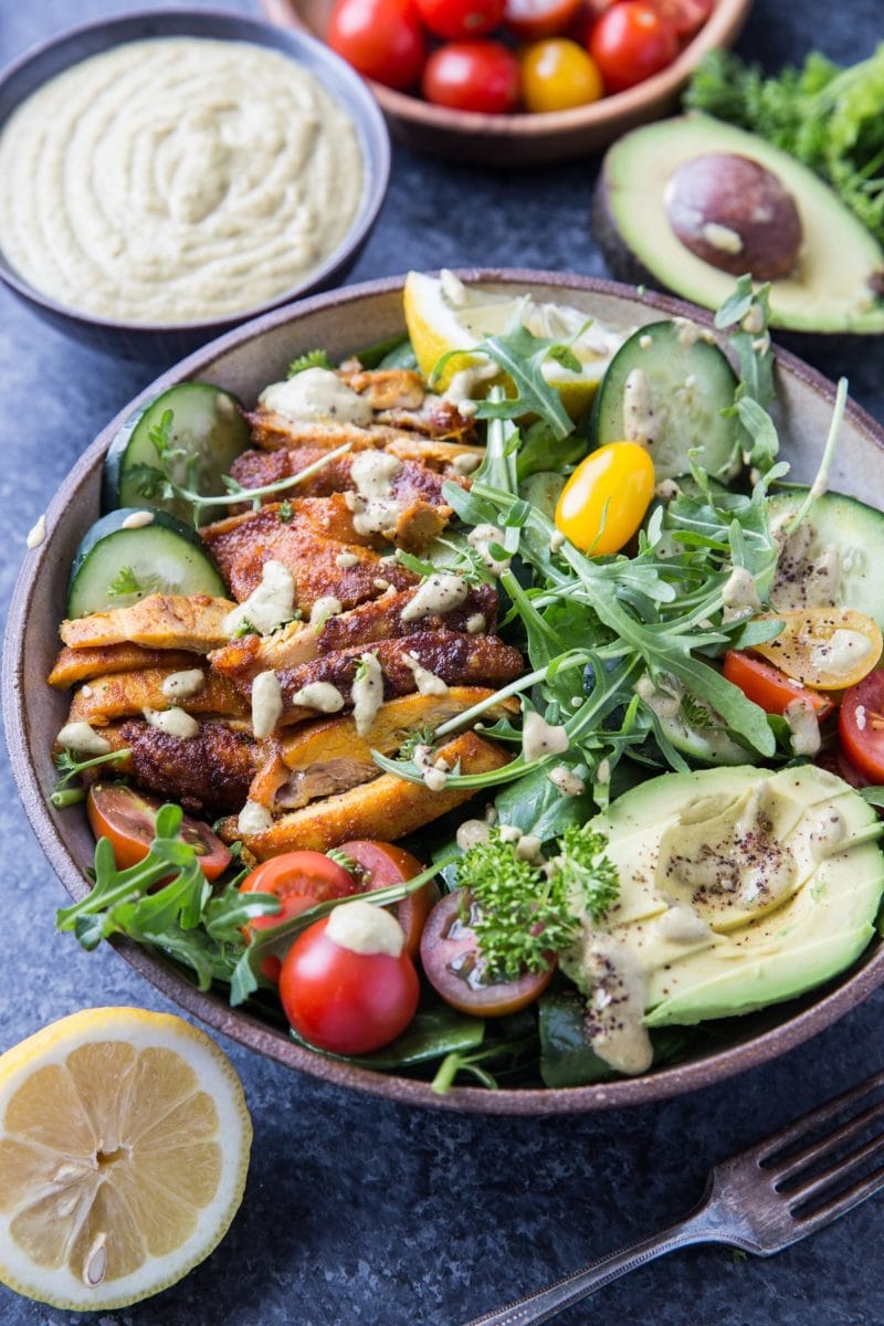 Angeled shot of a bowl of chicken salad with arugula, salad dressing and fresh tomatoes in the background and a fork up front.