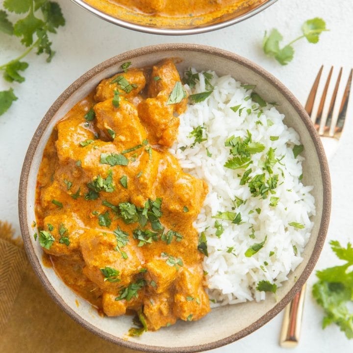Bowl of chicken tikka masala with rice and a serving dish of chicken tikka masala with a gold fork and a gold napkin.