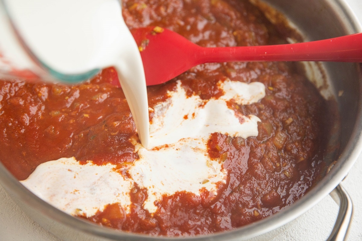 Skillet with spices, onions, tomato sauce and heavy cream being poured in.