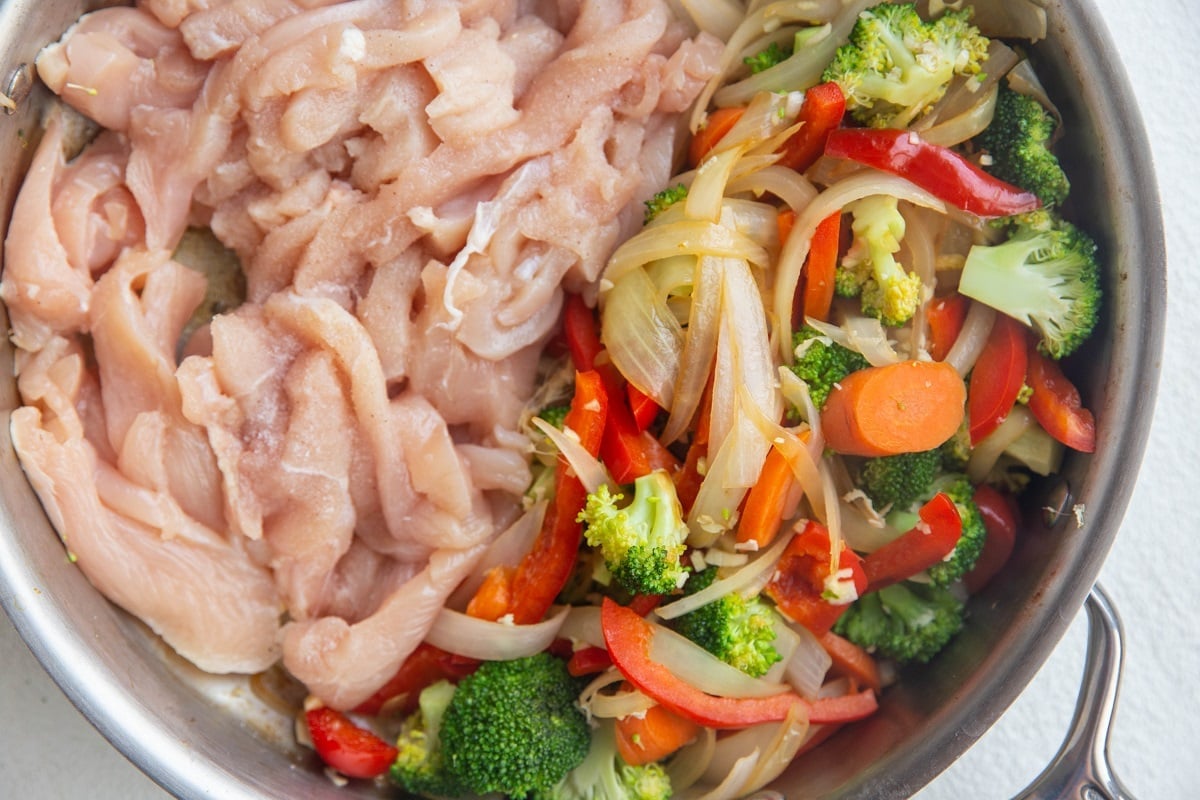 Stainless steel skillet with raw chicken breasts on half of the skillet and sauteed vegetables on half of the skillet. Browning the chicken.