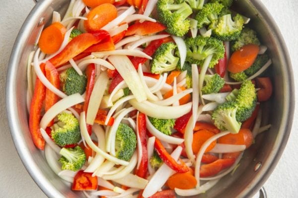 Onion, carrots, broccoli, and bell pepper cooking in a large stainless steel skillet.