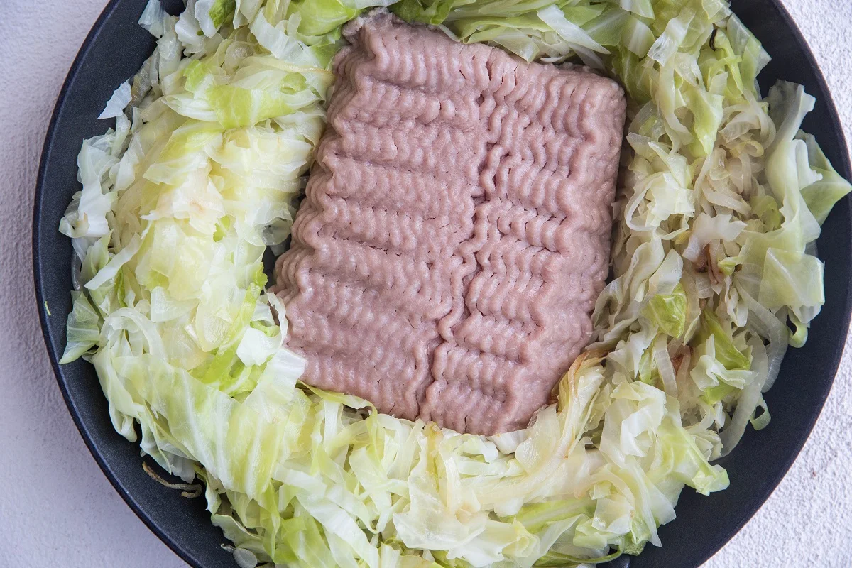Raw ground turkey in the center of a skillet with cooked cabbage and onions around it.
