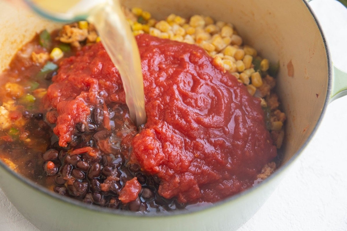 Crushed tomatoes on top of black beans, corn, turkey, onion and peppers, with chicken broth being poured into the pot.