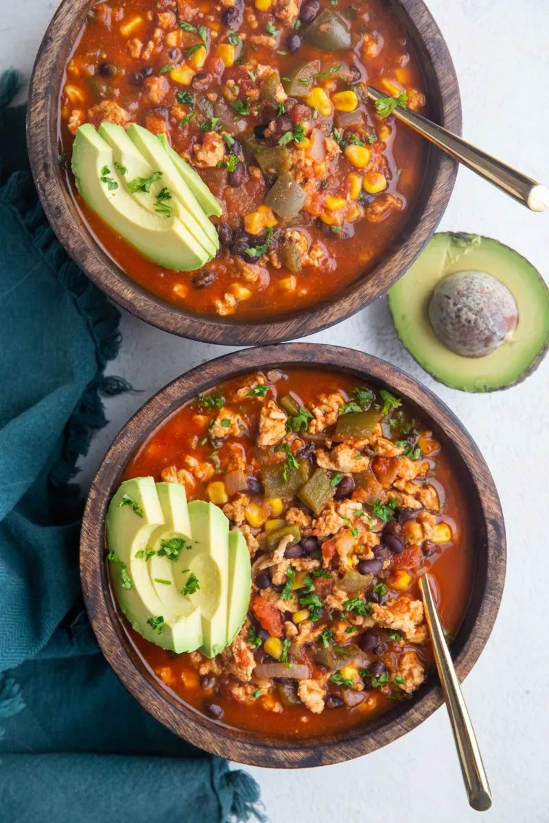 Two wooden bowls of taco soup with sliced avocado on top. Half of a fresh avocado to the side and a blue napkin on a white background.