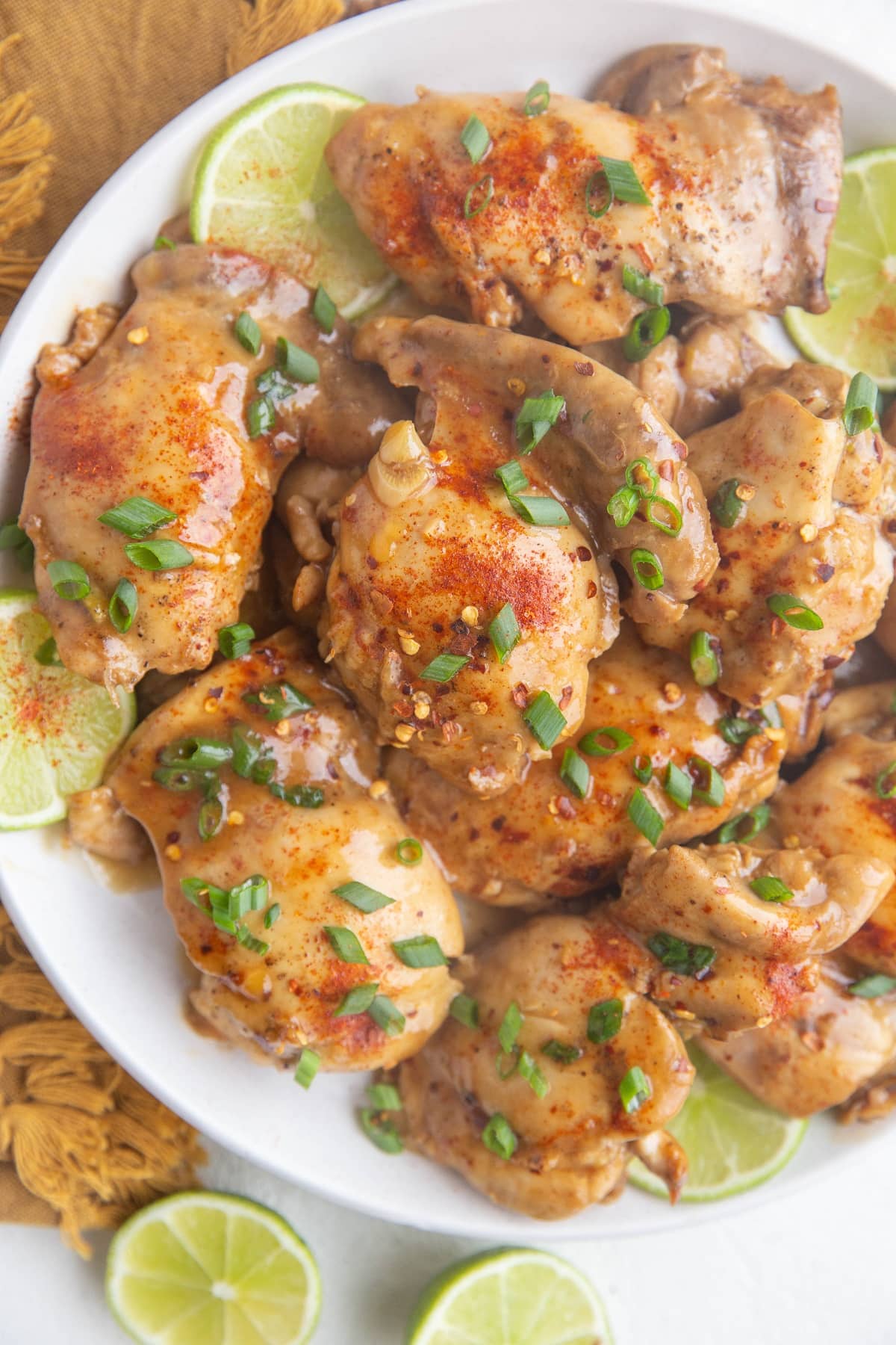 Top down white plate of cooked chicken thighs sprinkled with green onions and slices of limes all around.