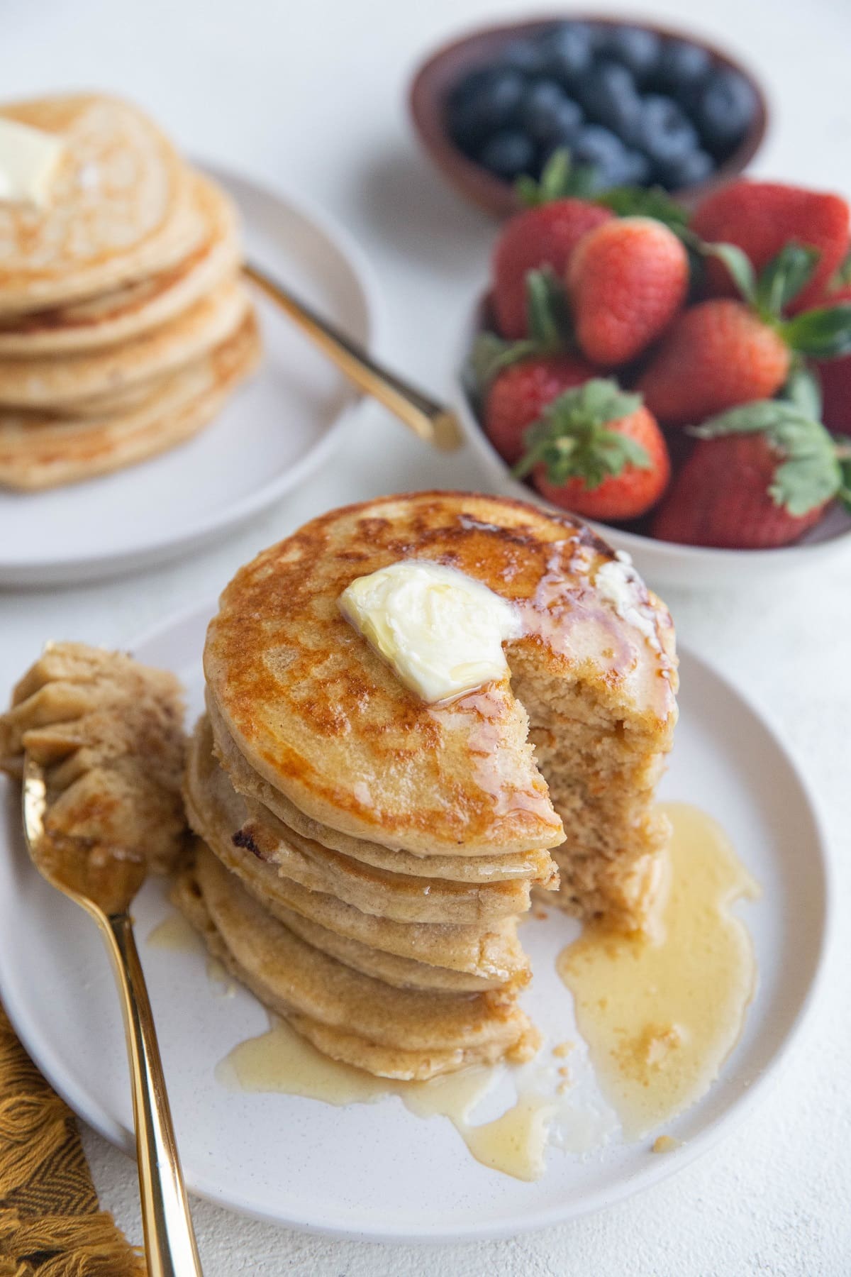 Plate of pancakes with a bite taken out with fresh strawberries and blueberries in the background and a second plate of pancakes.