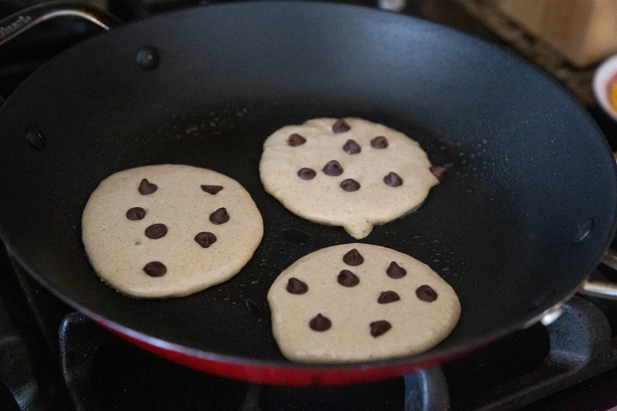 Three small pancakes cooking on a skillet with chocolate chips sprinkled on top.