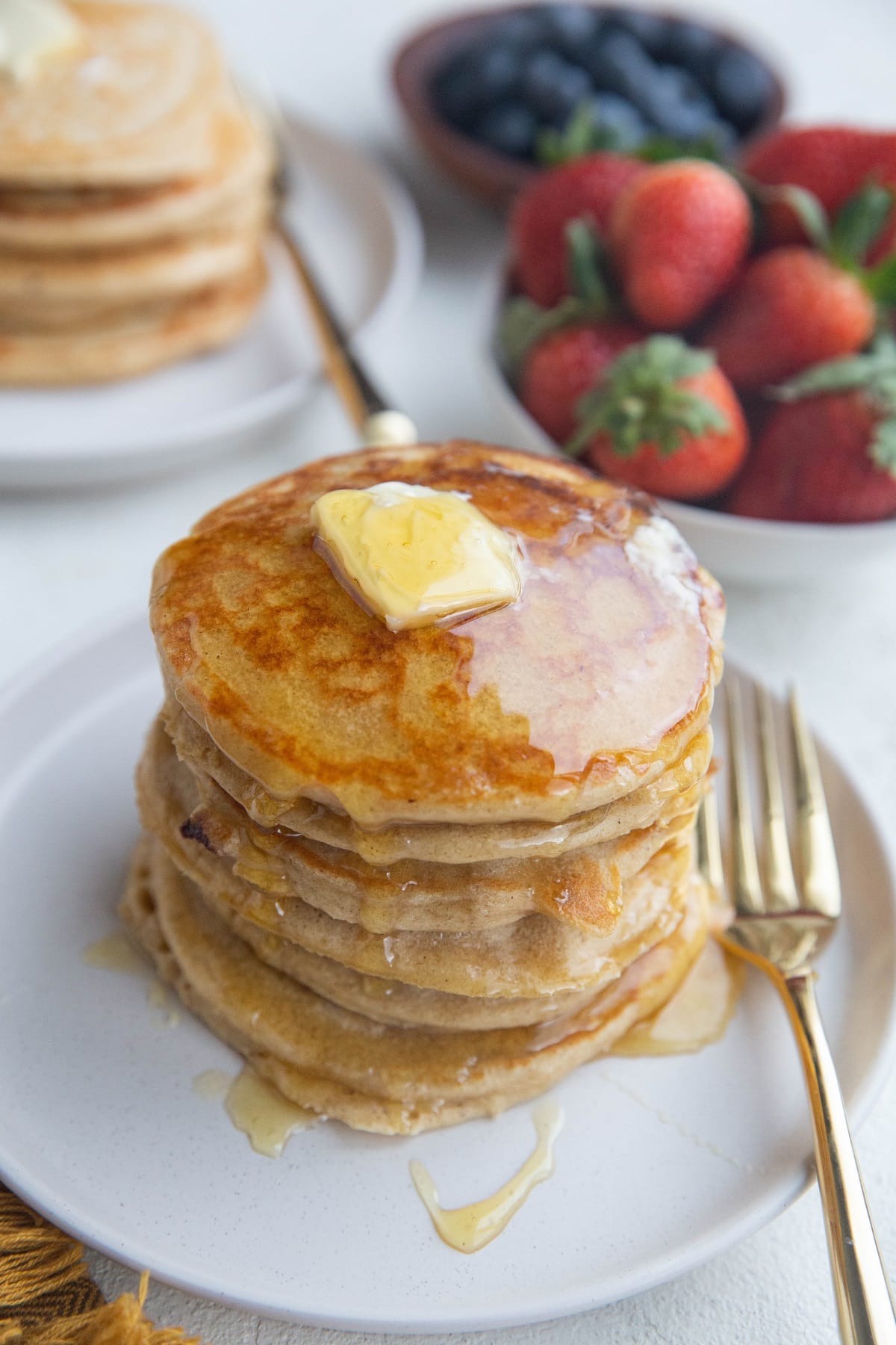 Stack of oatmeal protein pancakes on a white plate with butter and honey on top. Fresh berries in the background and a second plate of pancakes in the background.