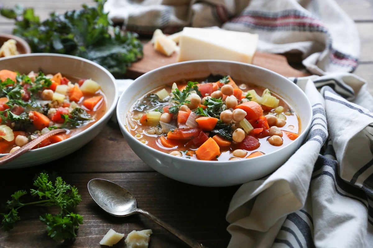 Two bowls of minestrone soup with napkins to the side and a cutting board with parmesan cheese in the background.