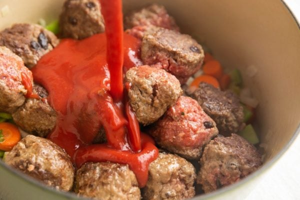 Tomato sauce being poured into a pot with veggies and meatballs.