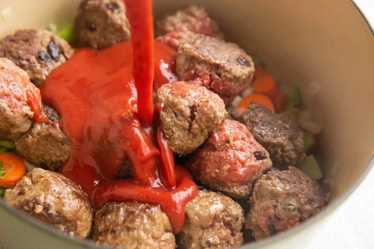Tomato sauce being poured into a pot with veggies and meatballs.