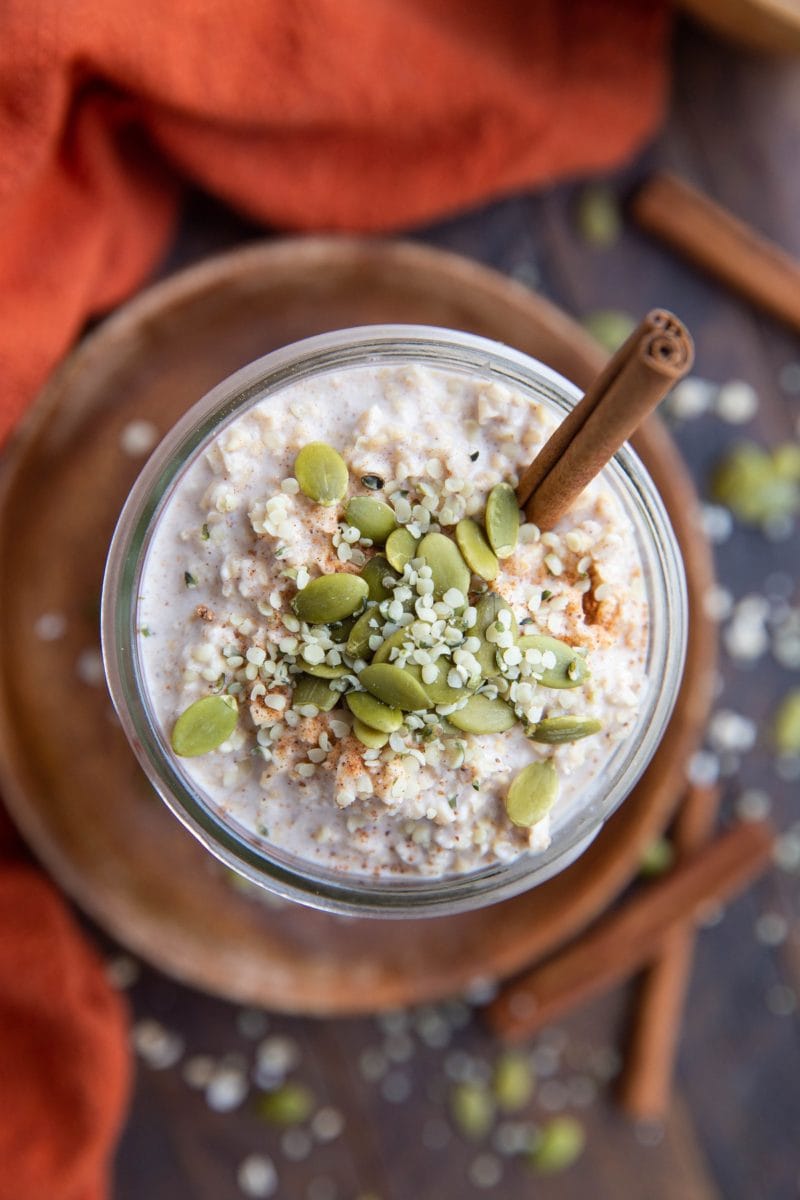 Top down photo of a jar of overnight oats with pumpkin seeds, hemp seeds and a cinnamon stick. Red napkin to the side with oats and pumpkin seeds sprinkled around.