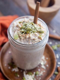 Jar full of overnight oatmeal sitting on a wooden plate on a wooden backdrop with oats and seeds all around as well as a red napkin.