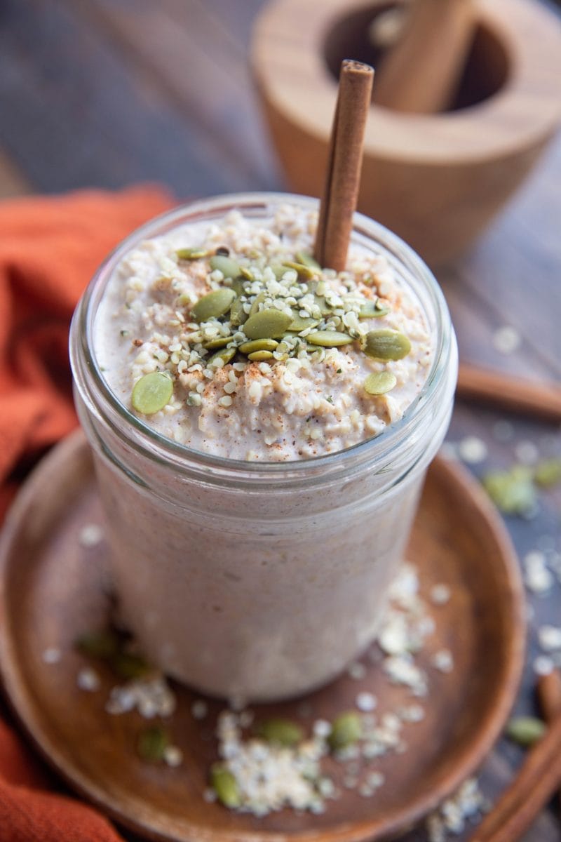 Jar full of overnight oatmeal sitting on a wooden plate on a wooden backdrop with oats and seeds all around as well as a red napkin.