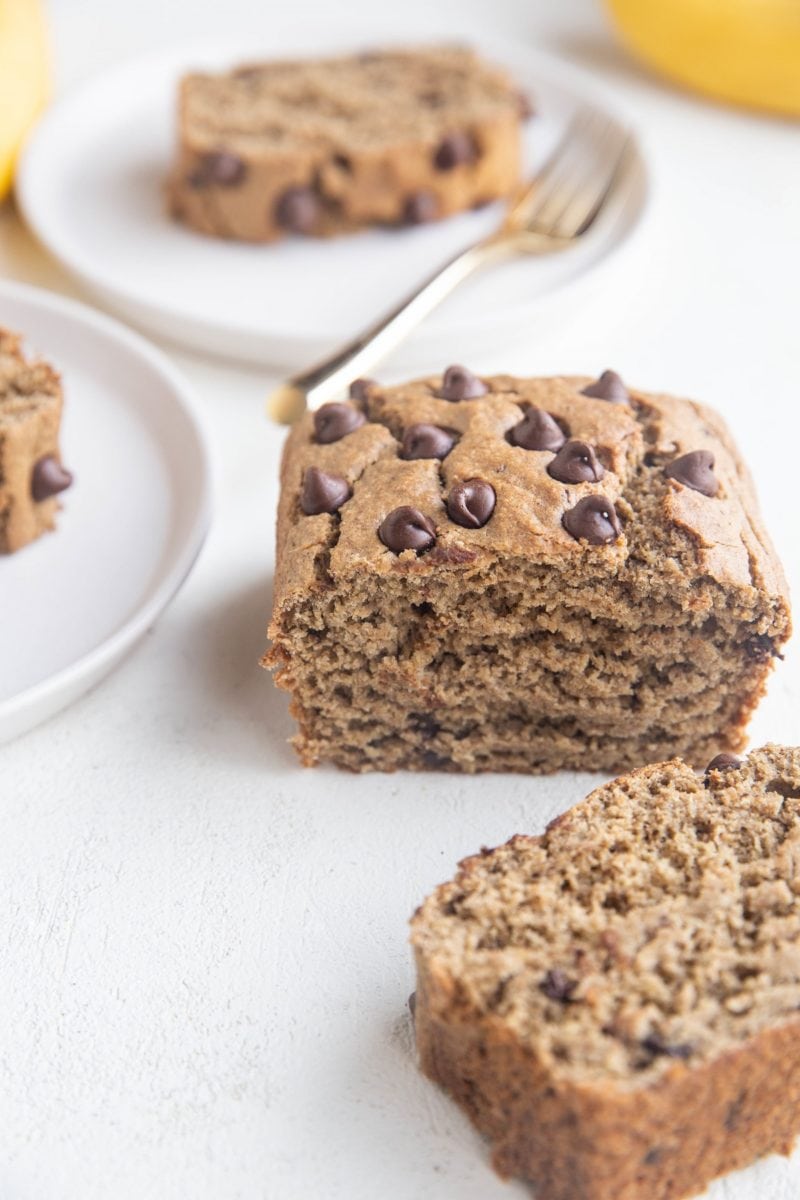 Loaf of banana bread on a white background with two plates of slices of banana bread in the background.