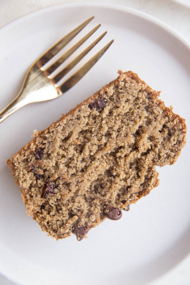 One large slice of banana bread on a white plate with a gold fork next to it, ready to serve.