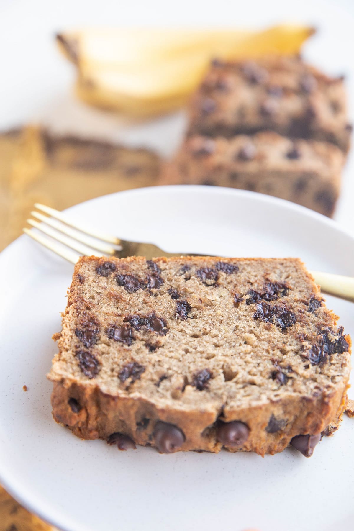 Big thick slice of banana bread on a white plate with a gold fork and the rest of the loaf of banana bread in the background.