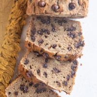 Loaf of grain-free banana bread cut into slices on a white backdrop with a gold napkin to the side.