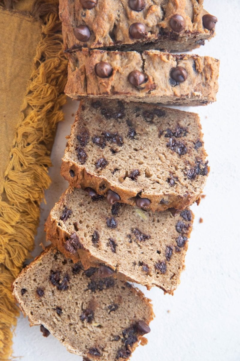 Loaf of grain-free banana bread cut into slices on a white backdrop with a gold napkin to the side.