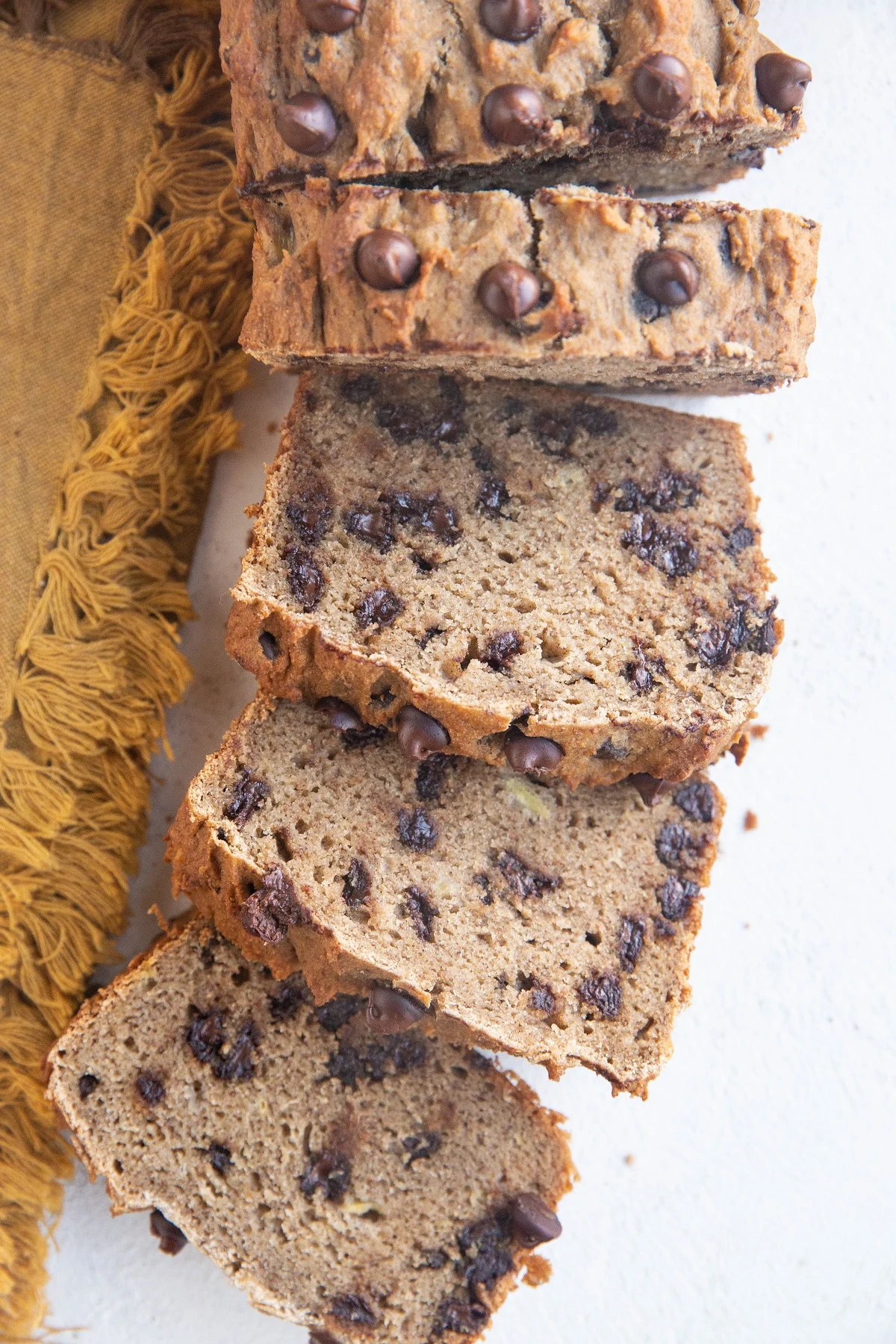 Loaf of grain-free banana bread cut into slices on a white backdrop with a gold napkin to the side.
