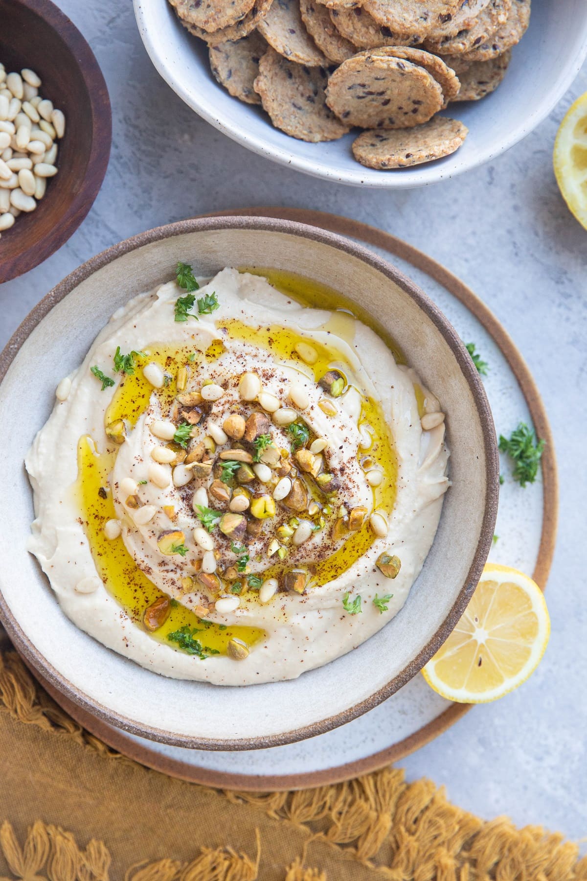 Bowl of hummus with pine nuts, pistachios and oil on top with a bowl of crackers to the side.