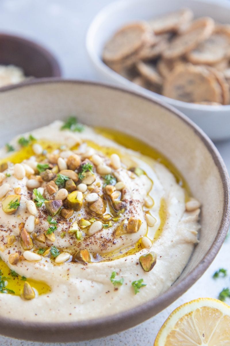 Big bowl of hummus with pine nuts and pistachios sprinkled on top and a drizzle of olive oil.