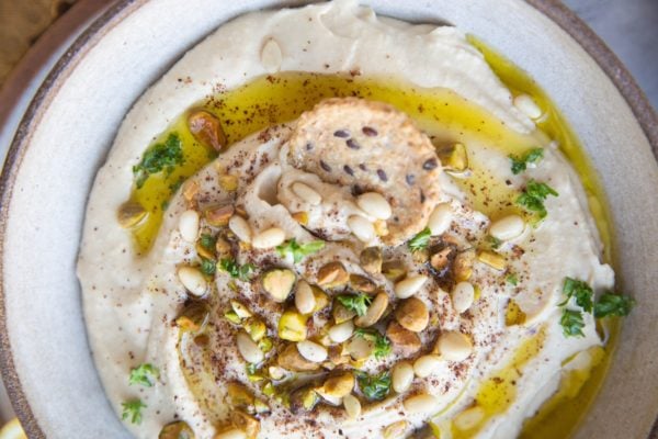 Bowl of hummus with nuts sprinkled on top and a cracker taking a bite out of the bowl.