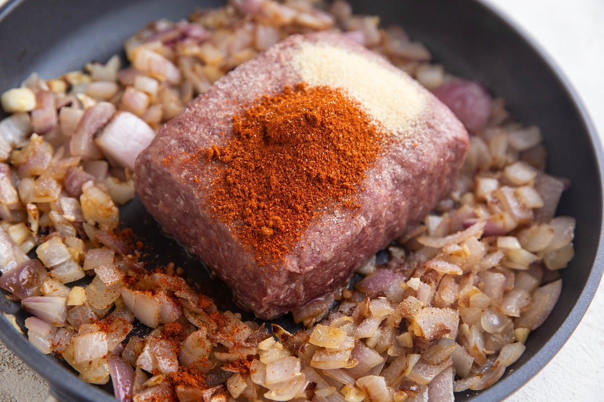 Ground beef in a skillet with chili powder and garlic powder sprinkled on top.