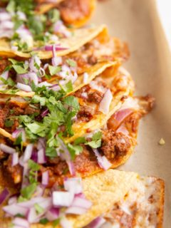 Pile of ground beef and cheese tacos laying on top of each other with cilantro and red onion sprinkled on top.