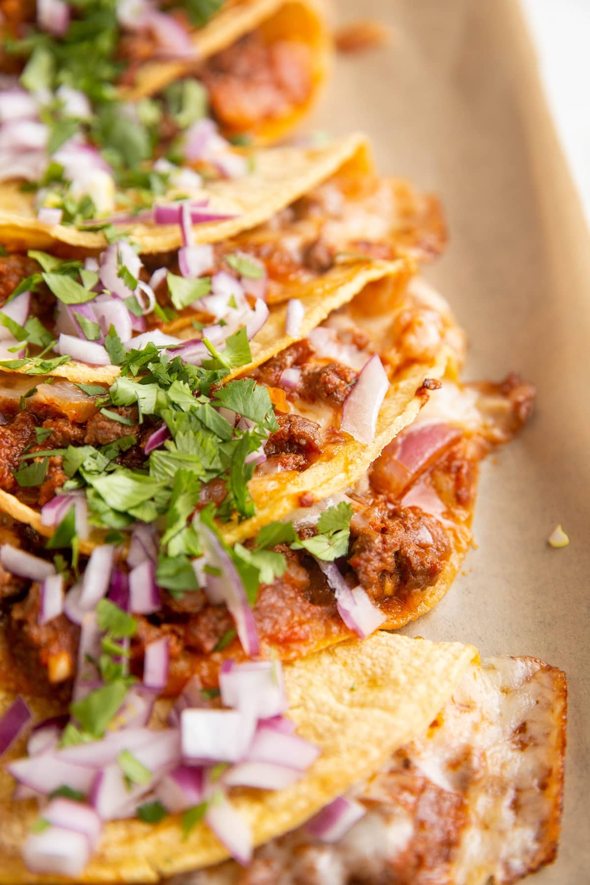 Pile of ground beef and cheese tacos laying on top of each other with cilantro and red onion sprinkled on top.