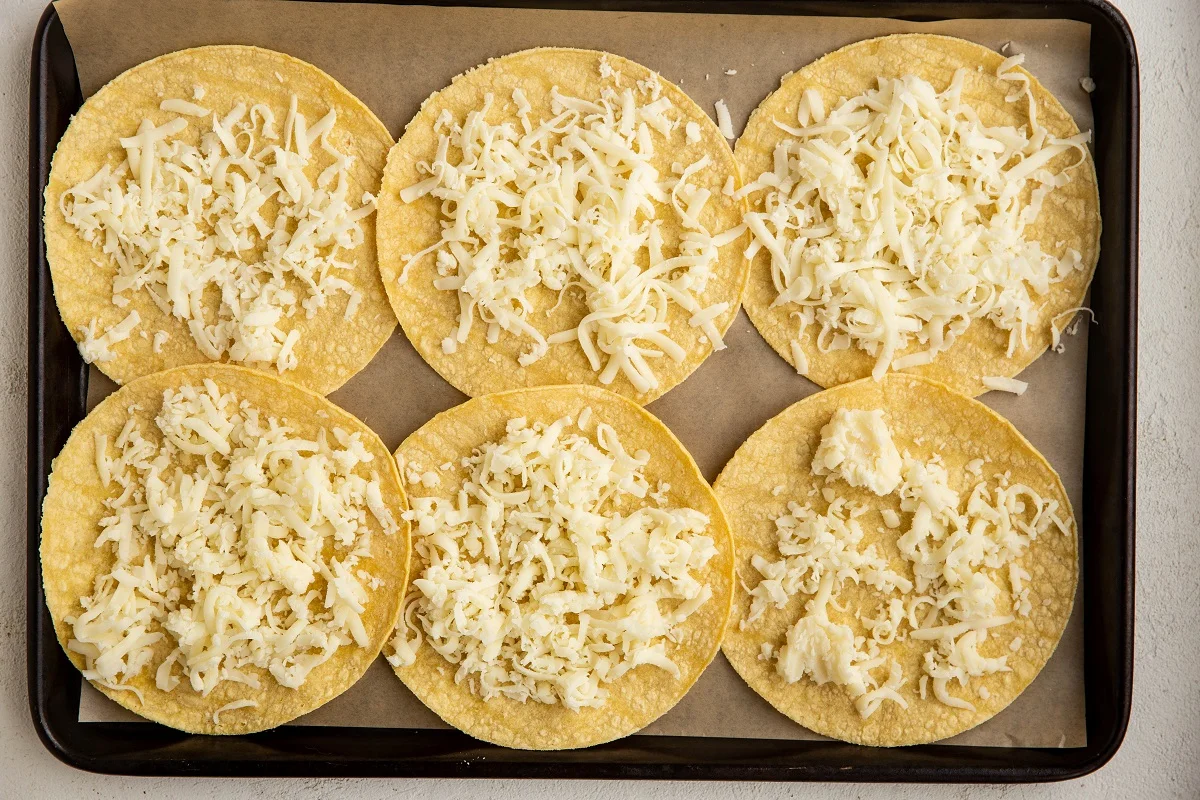Six tortillas on a parchment-lined baking sheet with grated cheese sprinkled on top of the tortillas, ready to go into the oven.