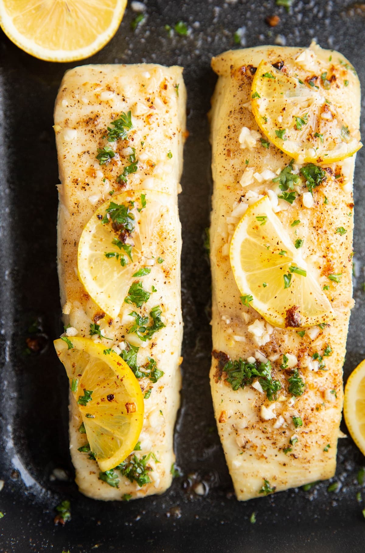 Two baked halibut filets in a black baking dish with slices of lemon on top.