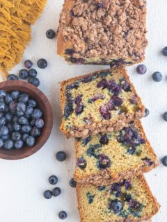 Cut up loaf of blueberry bread on a white background with fresh blueberries all around.