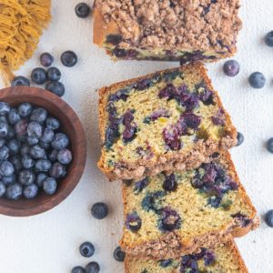 Cut up loaf of blueberry bread on a white background with fresh blueberries all around.