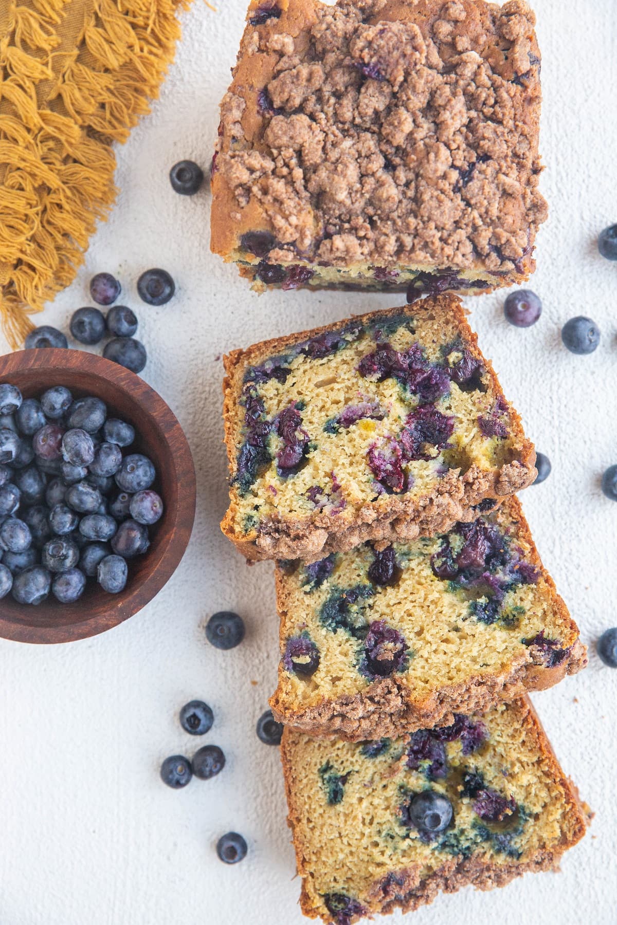 Cut up loaf of blueberry bread on a white background with fresh blueberries all around.