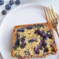 Slice of blueberry bread on a white plate with a gold fork and a bowl of fresh blueberries to the side.