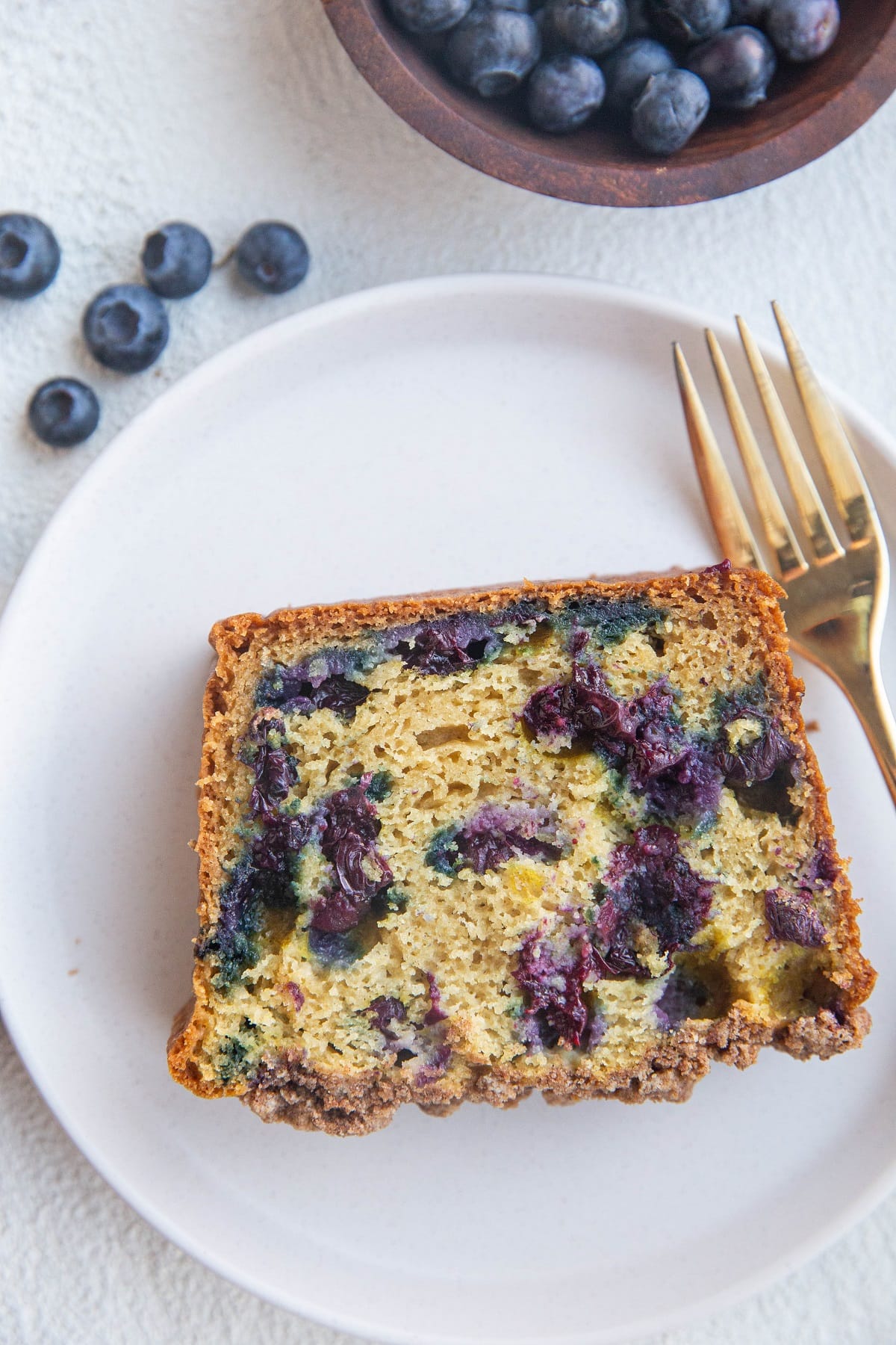 Slice of blueberry bread on a white plate with a gold fork and a bowl of fresh blueberries to the side.