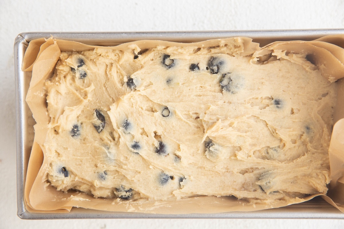 Blueberry muffin batter spread into an even layer in a prepared loaf pan, ready to go into the oven.