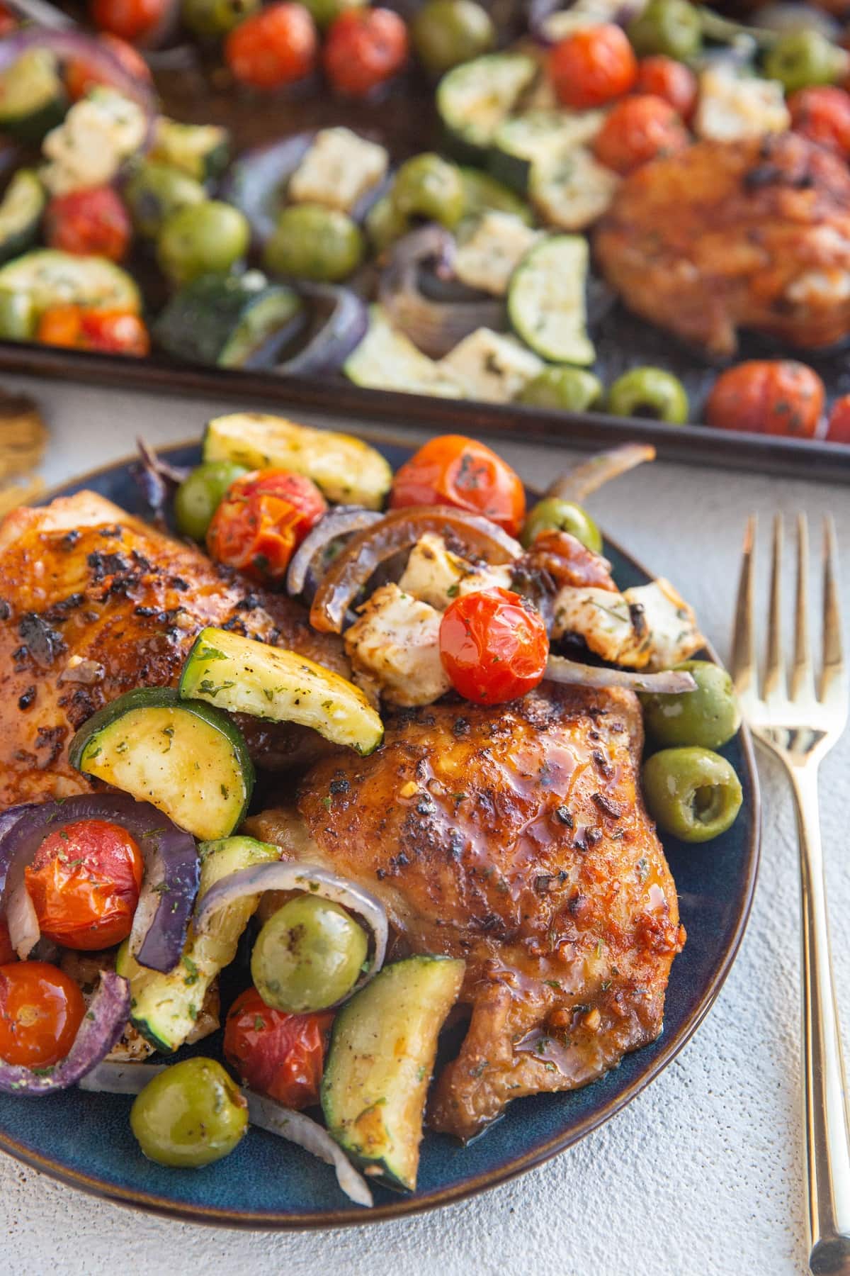 Plate with two chicken thighs and lots of vegetables on top with a sheet pan of chicken and vegetables in the background.