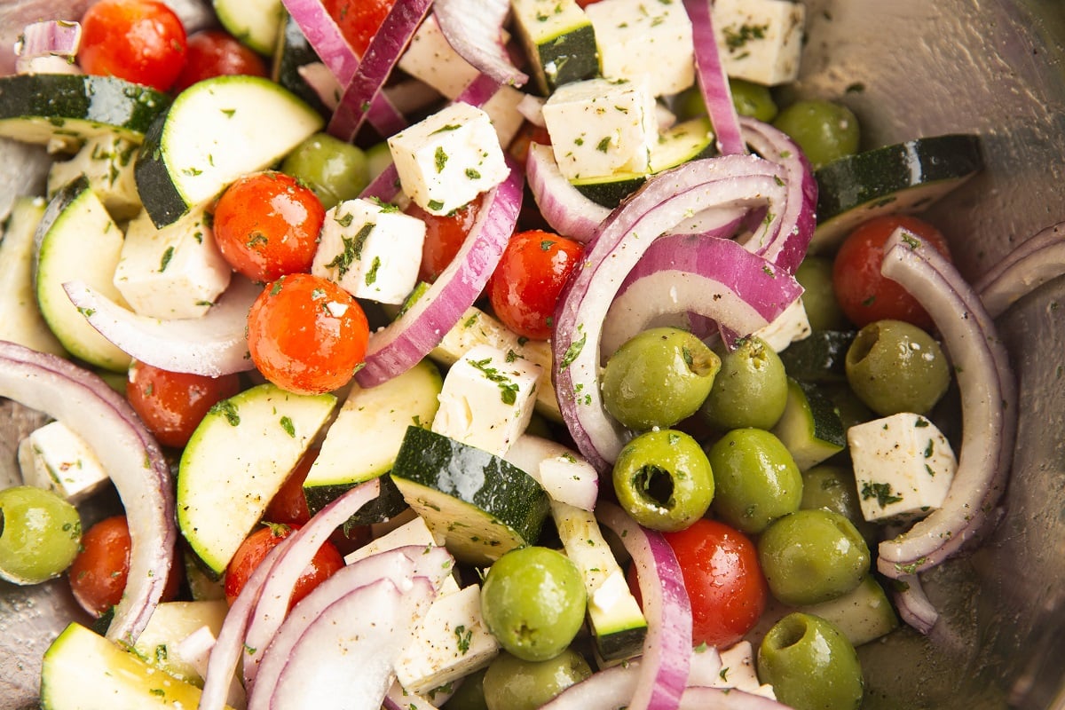tomatoes, onions, olives, feta cheese, and zucchini in a large mixing bowl with oil and dried herbs.