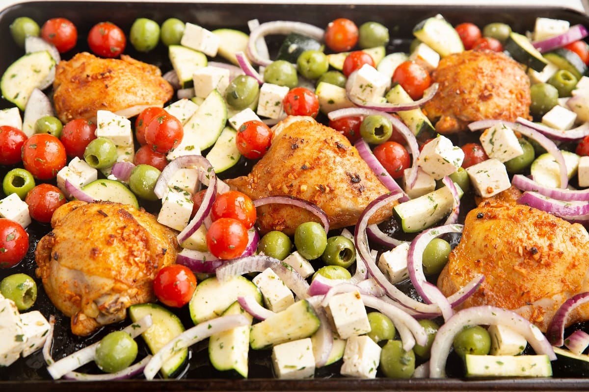 Chicken thighs on a baking sheet with cherry tomatoes, feta cheese, zucchini, onions, and olives, ready to go into the oven.