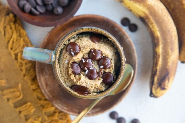 Finished banana bread mug cake in a mug with ripe bananas, a spoon, and chocolate chips to the side. Ready to serve.