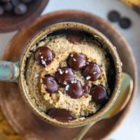 Mug full of banana mug cake sitting on a wooden plate with chocolate chips and ripe bananas to the side and a gold spoon.