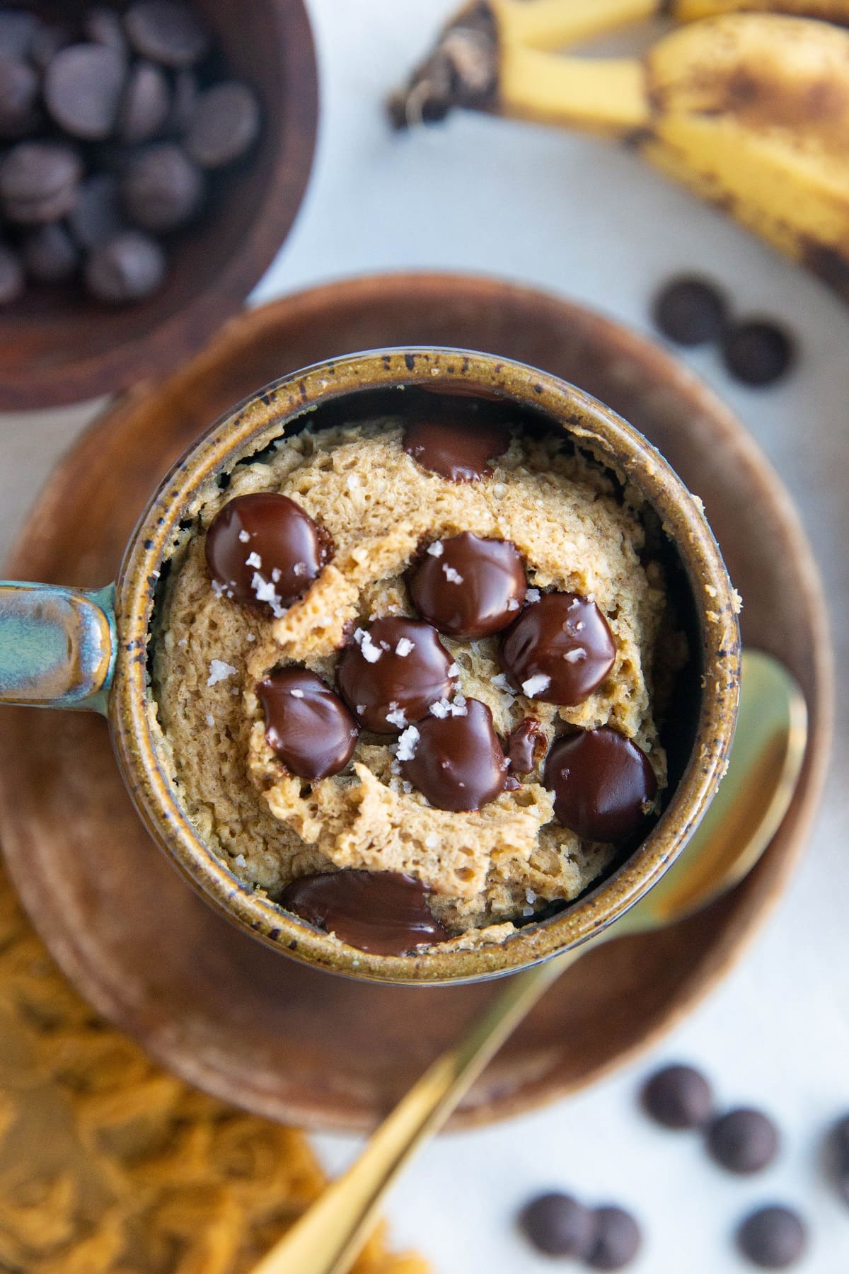 Mug full of banana mug cake sitting on a wooden plate with chocolate chips and ripe bananas to the side and a gold spoon.