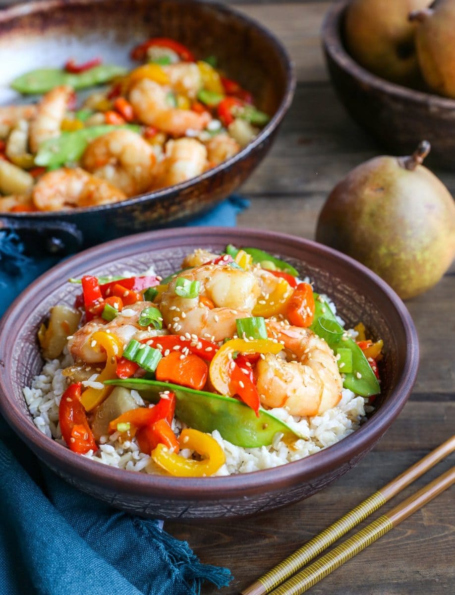 Bowl of shrimp stir fry with chopsticks and a blue napkin next to the bowl and a skillet with more shrimp stir fry in the background, ready to eat.