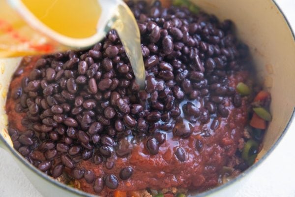 Ground beef, peppers, and onions in a pot with crushed tomatoes, black beans, and broth being poured in.