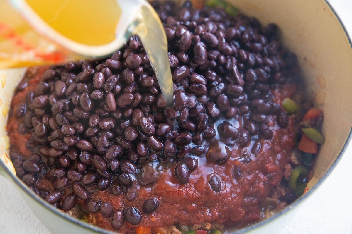 Ground beef, peppers, and onions in a pot with crushed tomatoes, black beans, and broth being poured in.